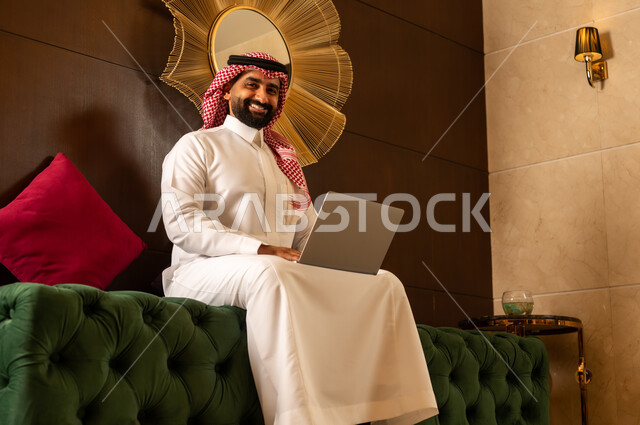 مفهوم اداره الاعمال عن بعد, استخدام التقنيات الحديثه في التواصل مع الاهل والاصدقاء وانجاز الاعمال,  الجلوس في إحدى الفنادق الراقية، رجل عربي خليجي سعودي يرتدي الشماغ والثوب التقليدي يجلس في لوبي الفندق ويعمل على جهاز الحاسوب المحمول