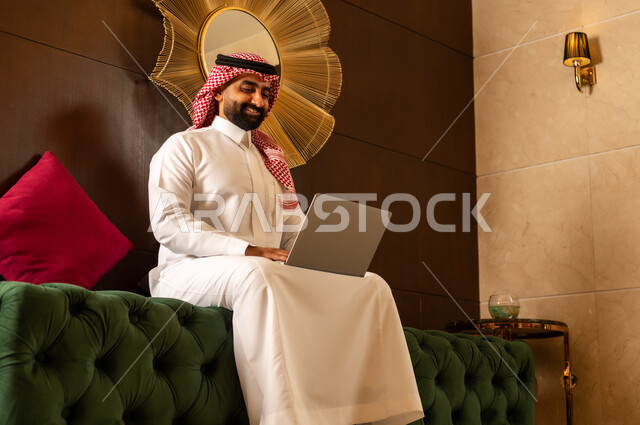 مفهوم اداره الاعمال عن بعد, استخدام التقنيات الحديثه في التواصل مع الاهل والاصدقاء وانجاز الاعمال,  الجلوس في إحدى الفنادق الراقية، رجل عربي خليجي سعودي يرتدي الشماغ والثوب التقليدي يجلس في لوبي الفندق ويعمل على جهاز الحاسوب المحمول