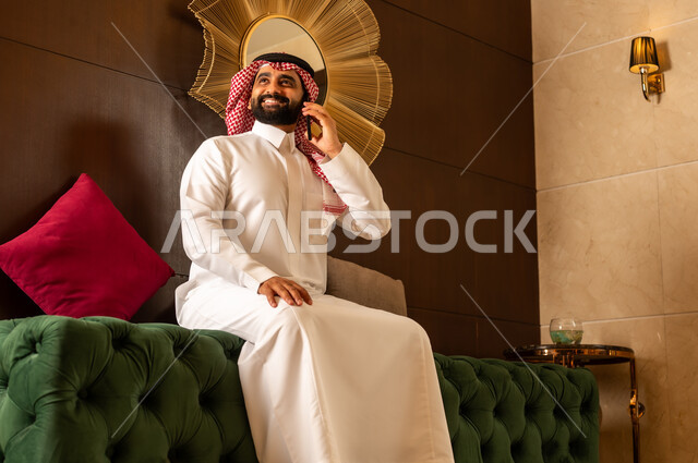 النظر إلى شيء ما أثناء إجراء مكالمة هاتفية عبر الجوال، الجلوس في لوبي إحدى الفنادق الراقية، استخدام جهاز تكنولوجي حديث ومتطور، رجل عربي خليجي سعودي يرتدي الشماغ والثوب التقليدي يقوم بالتحدث على الهاتف المحمول