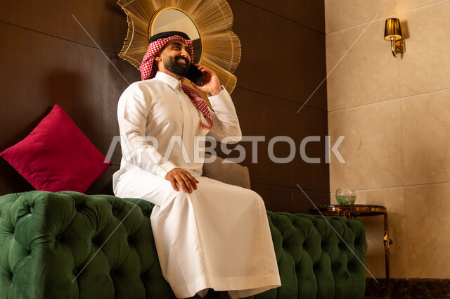 النظر إلى شيء ما أثناء إجراء مكالمة هاتفية عبر الجوال، الجلوس في لوبي إحدى الفنادق الراقية، استخدام جهاز تكنولوجي حديث ومتطور، رجل عربي خليجي سعودي يرتدي الشماغ والثوب التقليدي يقوم بالتحدث على الهاتف المحمول