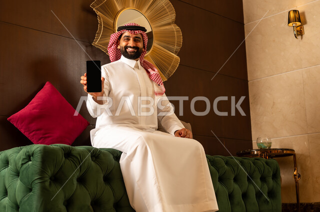 عرش شاشة سوداء فارغة على الجوال، الجلوس في لوبي إحدى الفنادق الراقية، استخدام جهاز تكنولوجي حديث ومتطور، رجل عربي خليجي سعودي يرتدي الشماغ والثوب التقليدي يقوم باستخدام الهاتف المحمول