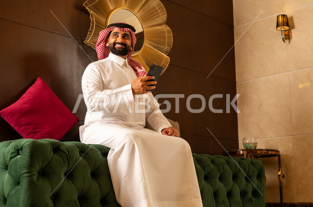 النظر إلى شيء ما أثناء العمل على الجوال، الجلوس في لوبي إحدى الفنادق الراقية، استخدام جهاز تكنولوجي حديث ومتطور، رجل عربي خليجي سعودي يرتدي الشماغ والثوب التقليدي يقوم باستخدام الهاتف المحمول