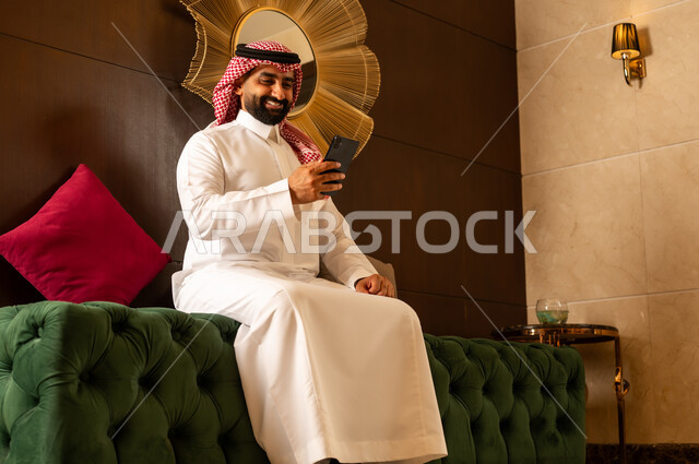 تعابير الاندماج بالعمل على الجوال، الجلوس في لوبي إحدى الفنادق الراقية، استخدام جهاز تكنولوجي حديث ومتطور، رجل عربي خليجي سعودي يرتدي الشماغ والثوب التقليدي يقوم باستخدام الهاتف المحمول