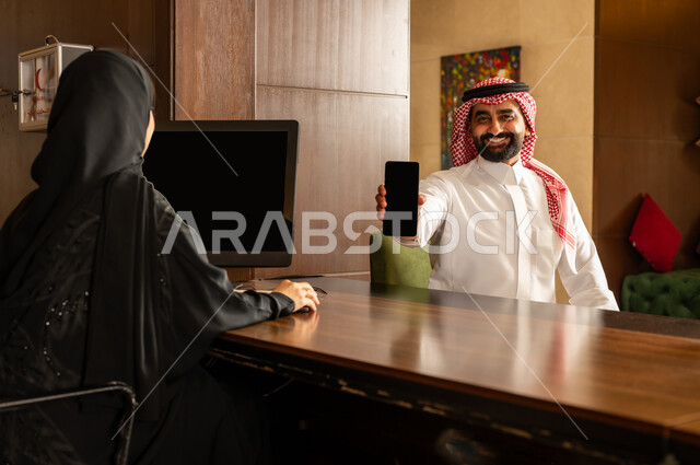 الدفع عن طريق بطاقة الائتمان البنكية عبر الهاتف المحمول, قضاء أوقات ممتعة في العطلة الصيفية, القيام بإجراءات الحجز والدخول, منتجعات وفنادق المملكة العربية السعودية، رجل عربي خليجي سعودي يرتدي الثوب التقليدي والشماغ يقوم بالدفع في مكتب الاستقبال للموظفة