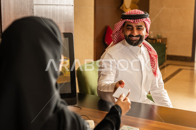 الدفع عن طريق بطاقة الائتمان البنكية عبر الهاتف المحمول, قضاء أوقات ممتعة في العطلة الصيفية, القيام بإجراءات الحجز والدخول, منتجعات وفنادق المملكة العربية السعودية، رجل عربي خليجي سعودي يرتدي الثوب التقليدي والشماغ يقوم بالدفع في مكتب الاستقبال للموظفة