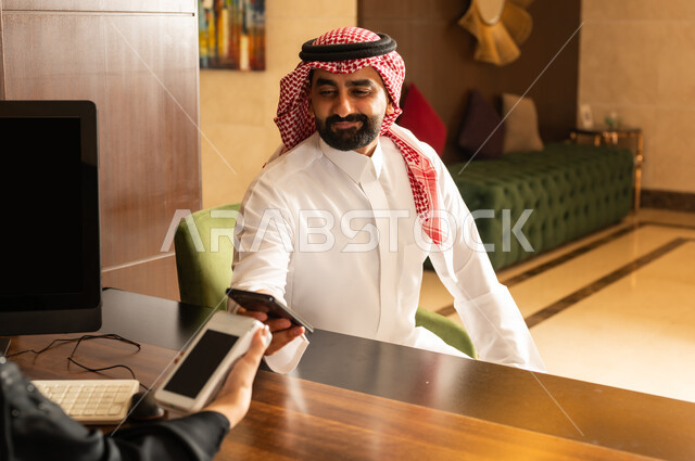 Paying by credit card via mobile phone, having a good time during the summer vacation, making reservations and entering, resorts and hotels in the Kingdom of Saudi Arabia, a Saudi Arabian Gulf man wearing a traditional thobe and shemagh paying at the reception desk to the employee