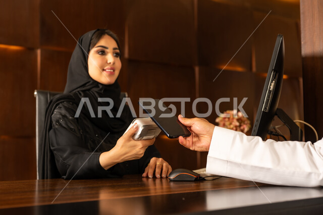 الدفع عن طريق بطاقة الائتمان البنكية عبر الهاتف المحمول, قضاء أوقات ممتعة في العطلة الصيفية, القيام بإجراءات الحجز والدخول, منتجعات وفنادق المملكة العربية السعودية، رجل عربي خليجي سعودي يرتدي الثوب التقليدي والشماغ يقوم بالدفع في مكتب الاستقبال للموظفة