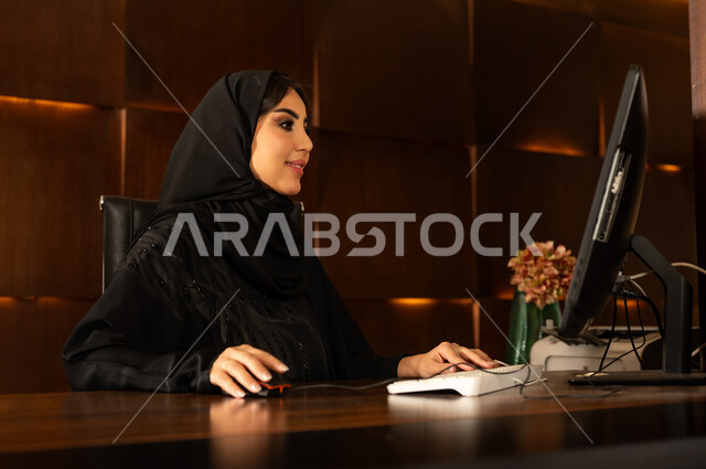 بيئة العمل في الفنادق السياحيه الراقية, الرد على استفسارات العملاء بتعابير البشاشة وحسن الاستقبال, مهن ووظائف نسائية, صورة مقربة من الجانب لشابة عربية خليجية سعودية ترتدي العباءة والحجاب تعمل بوظيفه حجز المواعيد