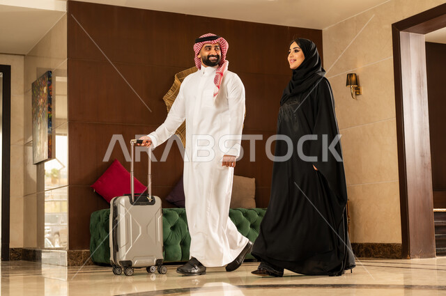 الإستعداد للسفر والسياحة وقضاء إجازة صيفية ممتعة، رجل عربي خليجي سعودي مبتسم يرتدي الشماغ والثوب التقليدي يقوم بجر حقيبة الامتعة برفقة زوجته في لوبي الفندق، رحلة عائلية سعيدة