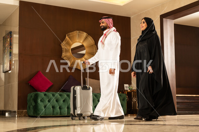 الإستعداد للسفر والسياحة وقضاء إجازة صيفية ممتعة، رجل عربي خليجي سعودي مبتسم يرتدي الشماغ والثوب التقليدي يقوم بجر حقيبة الامتعة برفقة زوجته في لوبي الفندق، رحلة عائلية سعيدة