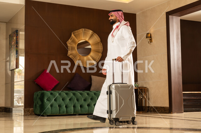 Preparing for a trip, the concept of tourism during summer vacation, a Saudi Arabian Gulf man wearing a traditional thobe and shemagh pulling a suitcase in the hotel lobby, expressions and gestures of happiness and pleasure, having a good time