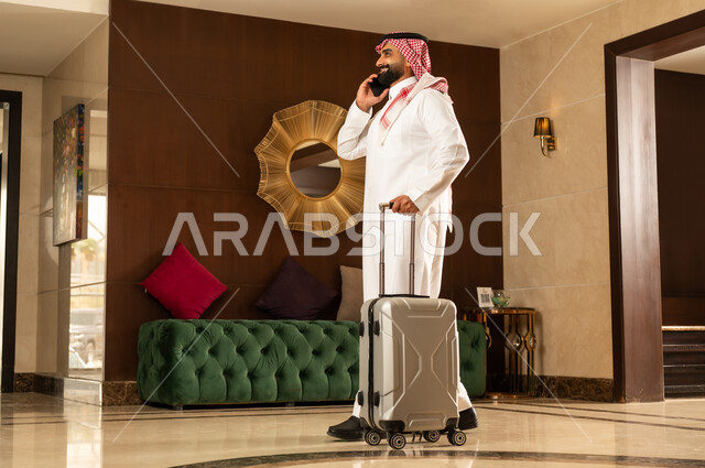 اجراء مكالمة هاتفية عبر الهاتف المحمول، الاستعداد للسفر في جولة سياحية، مفهوم السياحة في إجازة الصيف، رجل عربي سعودي خليجي يرتدي الثوب التقليدي والشماغ يقوم بجر حقيبة الامتعة في بهو الفندق، تعابير وايماءات السعادة والسرور، قضاء وقت ممتع