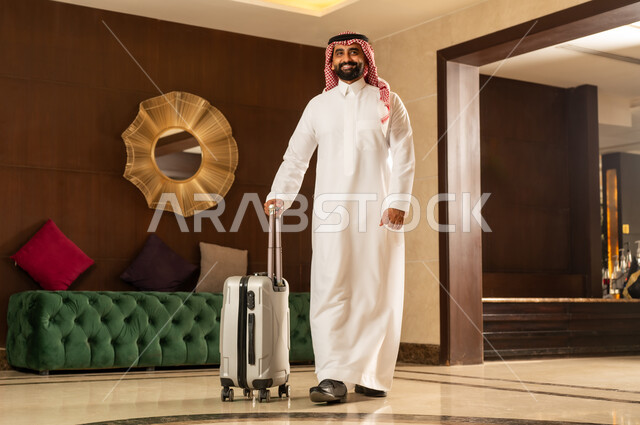 الاستعداد للسفر في جولة سياحية، مفهوم السياحة في إجازة الصيف، رجل عربي سعودي خليجي يرتدي الثوب التقليدي والشماغ يقوم بجر حقيبة الامتعة في بهو الفندق، تعابير وايماءات السعادة والسرور، قضاء وقت ممتع