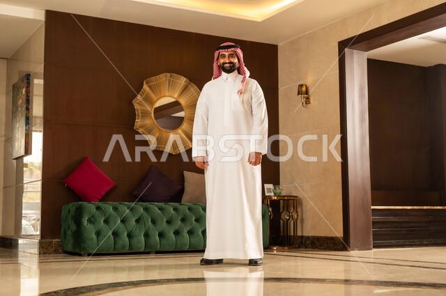 مفهوم الأناقة والموضة والاهتمام بالمظهر الخارجي، ايماءات الثقة بالنفس، رجل عربي سعودي خليجي يرتدي الثوب التقليدي والشماغ يقف باستقامة امام خلفيه ديكوريه حديثه بتعابير السرور السعادة