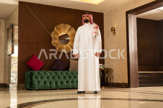 مفهوم الأناقة والموضة والاهتمام بالمظهر الخارجي، ايماءات الثقة بالنفس، رجل عربي سعودي خليجي يرتدي الثوب التقليدي والشماغ يقف باستقامة امام خلفيه ديكوريه حديثه بتعابير السرور السعادة