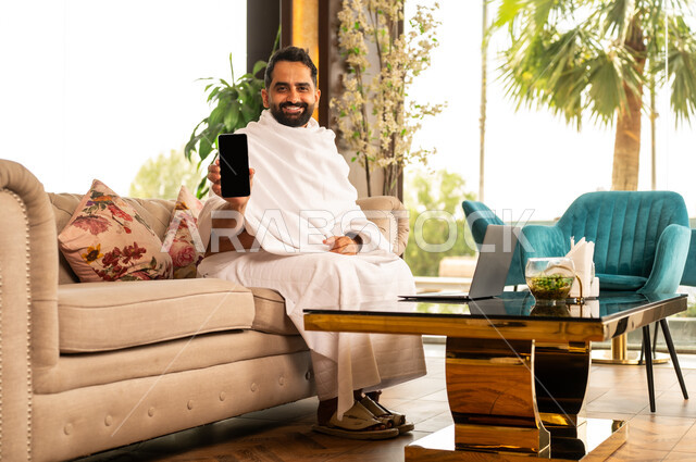 Preparing to perform Hajj and Umrah rituals, getting closer to Allah through good deeds, a smiling Saudi Arabian Gulf man wearing Ihram robe sitting on a comfortable sofa in the hotel lobby viewing a blank black screen on his mobile phone, performing religious rituals, the fifth pillar of Islam