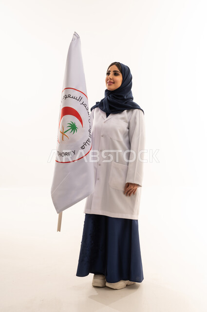 الإسهام في الخدمات الإنسانية والاجتماعية، تقديم الخدمة الطبية الإسعافية الطارئة، علم هيئة الهلال الأحمر السعودي، العمل في المجال الطبي، بورتريه لطبيبة سعودية عربية خليجية ترتدي المعطف الأبيض والحجاب تقف باستقامه بتعابير الثقة بالنفس، الجسم بالطول الكامل، خلفية بيضاء