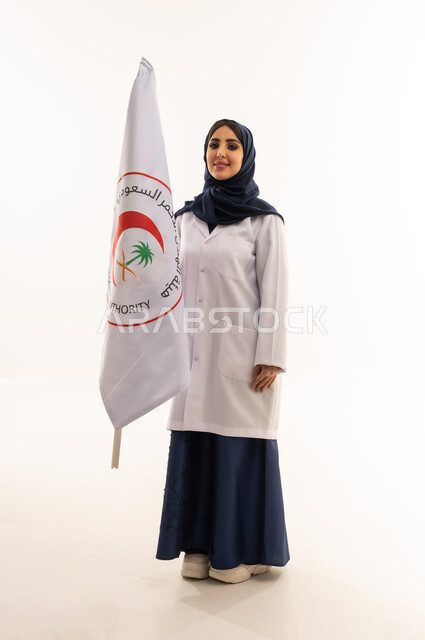 الإسهام في الخدمات الإنسانية والاجتماعية، تقديم الخدمة الطبية الإسعافية الطارئة، علم هيئة الهلال الأحمر السعودي، العمل في المجال الطبي، بورتريه لطبيبة سعودية عربية خليجية ترتدي المعطف الأبيض والحجاب تقف باستقامه بتعابير الثقة بالنفس، الجسم بالطول الكامل، خلفية بيضاء