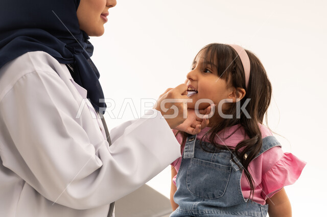 مهنة ووظيفية نسائية طبية، تعابير السرور واللطف، عيادة التمريض والتشخيص الطبي في السعودية، الحاجة إلى العلاج والمساعدة الصحية, بورتريه مقرب لطبيبة عربية خليجية سعودية محجبة ترتدي المعطف الابيض تقوم بقياس حرارة فتاة، خلفية بيضاء