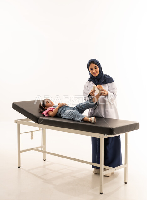 Extremity injuries, hospitals and treatment centers in Saudi Arabia, portrait of a veiled Saudi Arabian Gulf Arab female doctor wearing a medical coat wrapping a bandage on a little girl's foot, female medical profession and job, white background