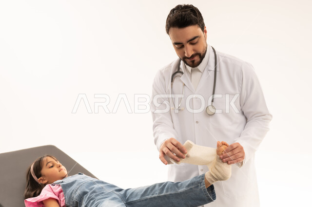 Extremity injuries, hospitals and treatment centers in Saudi Arabia, portrait of a Saudi Arabian Gulf doctor wearing a traditional thobe with a white coat wrapping a bandage on a little girl's foot, medical profession and occupation, white background
