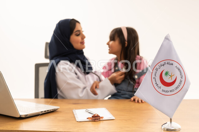 هيئة الهلال الأحمر السعودي، تقديم الخدمة الطبية الإسعافية الطارئة بالمملكة، العمل على الحاسوب المحمول في المجال الطبي والصحي، الإسهام في الخدمات الإنسانية والاجتماعية، بورتريه لطبيبة سعودية عربية خليجية ترتدي المعطف الأبيض والحجاب تقوم بفحص فتاة، خلفية بيضاء