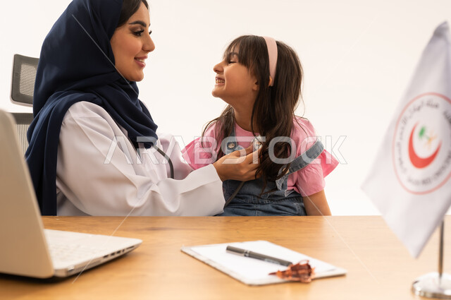 هيئة الهلال الأحمر السعودي، تقديم الخدمة الطبية الإسعافية الطارئة بالمملكة، العمل على الحاسوب المحمول في المجال الطبي والصحي، الإسهام في الخدمات الإنسانية والاجتماعية، بورتريه لطبيبة سعودية عربية خليجية ترتدي المعطف الأبيض والحجاب تقوم بفحص فتاة، خلفية بيضاء