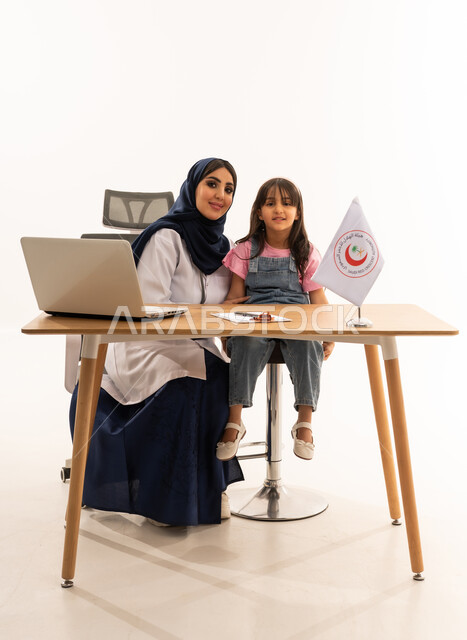 تقديم الخدمة الطبية الإسعافية الطارئة بالمملكة، العمل على الحاسوب المحمول في المجال الطبي والصحي، هيئة الهلال الأحمر السعودي، الإسهام في الخدمات الإنسانية والاجتماعية، بورتريه لطبيبة سعودية عربية خليجية ترتدي المعطف الأبيض والحجاب تقوم بفحص فتاة، خلفية بيضاء