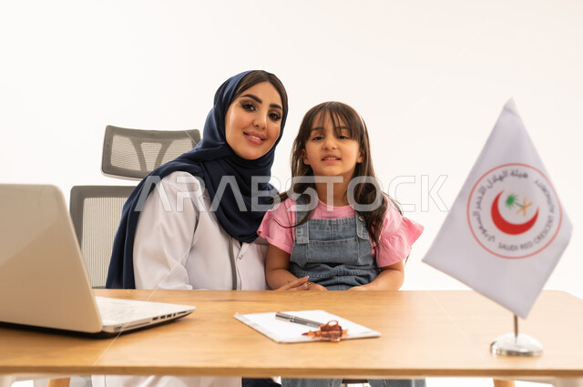 الإسهام في الخدمات الإنسانية والاجتماعية، هيئة الهلال الأحمر السعودي، تقديم الخدمة الطبية الإسعافية الطارئة بالمملكة، العمل على الحاسوب المحمول في المجال الطبي والصحي، بورتريه لطبيبة سعودية عربية خليجية ترتدي المعطف الأبيض والحجاب تقوم بملاطفة فتاة، خلفية بيضاء