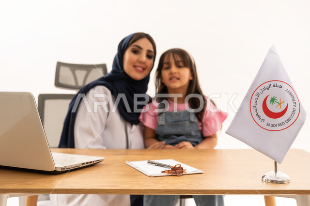 الإسهام في الخدمات الإنسانية والاجتماعية، هيئة الهلال الأحمر السعودي، تقديم الخدمة الطبية الإسعافية الطارئة بالمملكة، العمل على الحاسوب المحمول في المجال الطبي والصحي، بورتريه لطبيبة سعودية عربية خليجية ترتدي المعطف الأبيض والحجاب تقوم بملاطفة فتاة، خلفية بيضاء