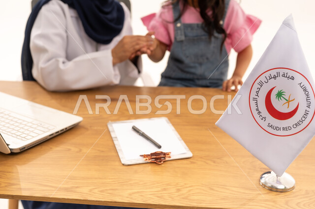 الإسهام في الخدمات الإنسانية والاجتماعية، هيئة الهلال الأحمر السعودي، تقديم الخدمة الطبية الإسعافية الطارئة بالمملكة، العمل على الحاسوب المحمول في المجال الطبي والصحي، بورتريه لطبيبة سعودية عربية خليجية ترتدي المعطف الأبيض والحجاب تقوم بملاطفة فتاة، خلفية بيضاء