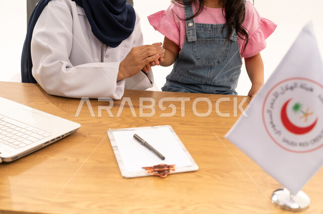 الإسهام في الخدمات الإنسانية والاجتماعية، هيئة الهلال الأحمر السعودي، تقديم الخدمة الطبية الإسعافية الطارئة بالمملكة، العمل على الحاسوب المحمول في المجال الطبي والصحي، بورتريه لطبيبة سعودية عربية خليجية ترتدي المعطف الأبيض والحجاب تقوم بملاطفة فتاة، خلفية بيضاء