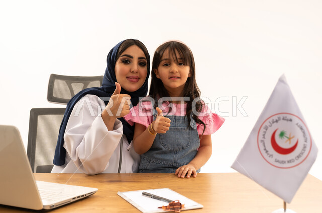 رفع اصبع الابهام بتعابير القبول والرضا، المساهمة في الخدمات الطبية الإسعافية الطارئة، العمل على الحاسوب المحمول في المجال الطبي والصحي، هيئة الهلال الأحمر السعودي، بورتريه لطبيبة سعودية عربية خليجية ترتدي المعطف الأبيض والحجاب تقوم بفحص فتاة، خلفية بيضاء