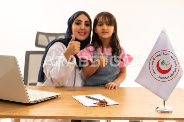 رفع اصبع الابهام بتعابير القبول والرضا، المساهمة في الخدمات الطبية الإسعافية الطارئة، العمل على الحاسوب المحمول في المجال الطبي والصحي، هيئة الهلال الأحمر السعودي، بورتريه لطبيبة سعودية عربية خليجية ترتدي المعطف الأبيض والحجاب تقوم بفحص فتاة، خلفية بيضاء