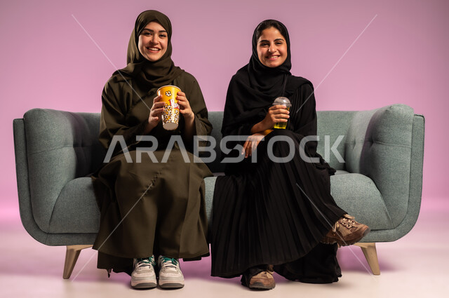 تناول الفوشار والعصير اثناء متابعة الأفلام المسلية، بورتريه لإمرأتين عربيتين خليجيتين سعوديتين ترتديان الحجاب والعباءة تجلسان على كنبة تقومان بأنشطة ترفيهيه في عطلة نهاية الإسبوع، قضاء وقت ممتع في السينما، خلفية ملونة