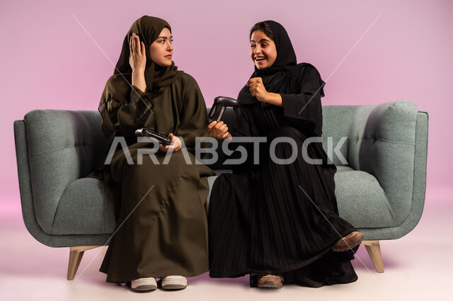 استخدام التقنية الحديثة المتطورة, قضاء وقت الفراغ بشكل ممتع مع الاصدقاء، بورتريه لامرأتين عربيتين خليجيتين سعوديتين ترتديان الحجاب والعباءة تجلسان على كنبة مريحة تمسكان بيدهما عصا التحكم بايماءات التفاعل والاندماج باللعب في العاب الفيديو, خلفية ملونة