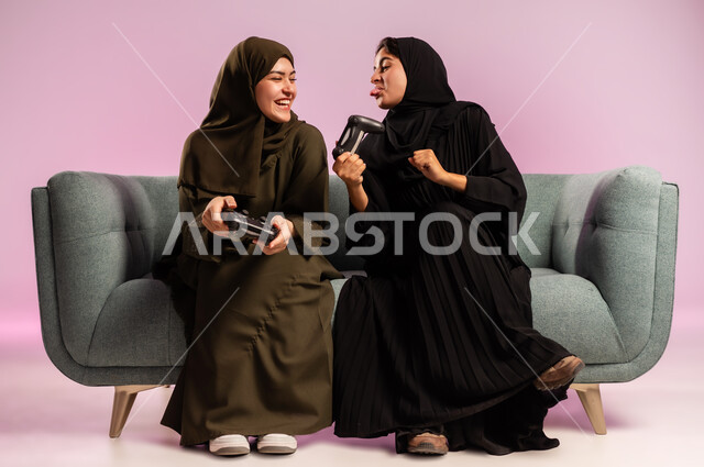 استخدام التقنية الحديثة المتطورة, قضاء وقت الفراغ بشكل ممتع مع الاصدقاء، بورتريه لامرأتين عربيتين خليجيتين سعوديتين ترتديان الحجاب والعباءة تجلسان على كنبة مريحة تمسكان بيدهما عصا التحكم بايماءات التفاعل والاندماج باللعب في العاب الفيديو, خلفية ملونة