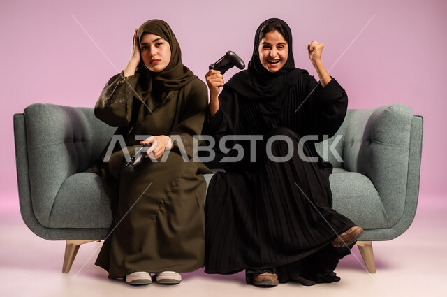 تعابير الخسارة والفشل، قضاء وقت الفراغ ممتع مع الاصدقاء، استخدام التقنية الحديثة المتطورة, بورتريه لامرأتين عربيتين خليجيتين سعوديتين ترتديان الحجاب والعباءة تجلسان على كنبة مريحة تمسكان بيدهما عصا التحكم بايماءات التفاعل والاندماج باللعب في العاب الفيديو, خلفية ملونة