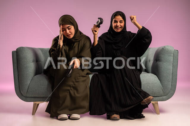 تعابير الخسارة والفشل، قضاء وقت الفراغ ممتع مع الاصدقاء، استخدام التقنية الحديثة المتطورة, بورتريه لامرأتين عربيتين خليجيتين سعوديتين ترتديان الحجاب والعباءة تجلسان على كنبة مريحة تمسكان بيدهما عصا التحكم بايماءات التفاعل والاندماج باللعب في العاب الفيديو, خلفية ملونة