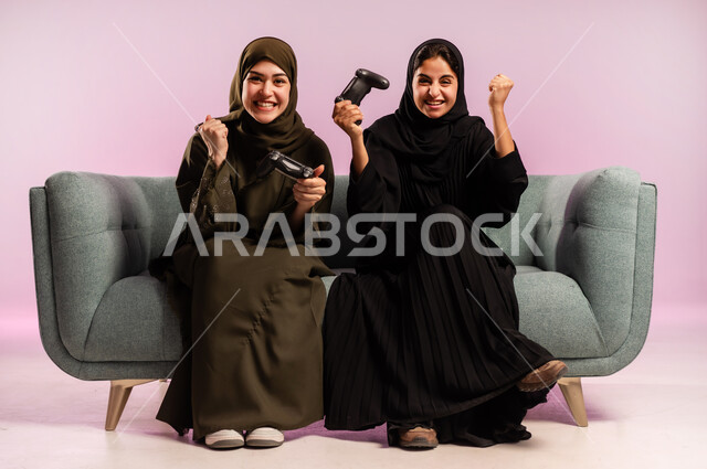 قضاء وقت الفراغ بشكل ممتع مع الاصدقاء، استخدام التقنية الحديثة المتطورة, بورتريه لامرأتين عربيتين خليجيتين سعوديتين ترتديان الحجاب والعباءة تجلسان على كنبة مريحة تمسكان بيدهما عصا التحكم بايماءات التفاعل والاندماج باللعب في العاب الفيديو, خلفية ملونة