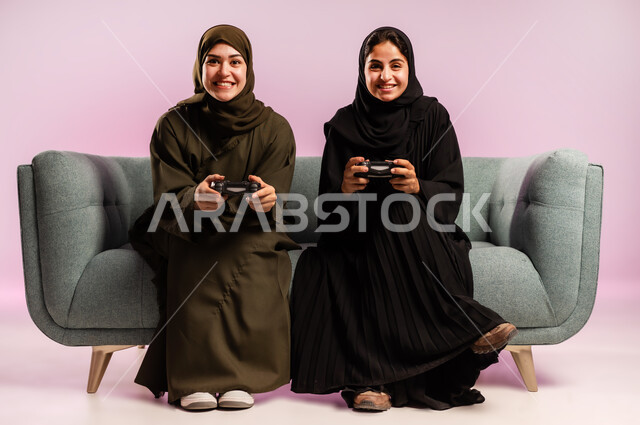 قضاء وقت الفراغ بشكل ممتع مع الاصدقاء، استخدام التقنية الحديثة المتطورة, بورتريه لامرأتين عربيتين خليجيتين سعوديتين ترتديان الحجاب والعباءة تجلسان على كنبة مريحة تمسكان بيدهما عصا التحكم بايماءات التفاعل والاندماج باللعب في العاب الفيديو, خلفية ملونة