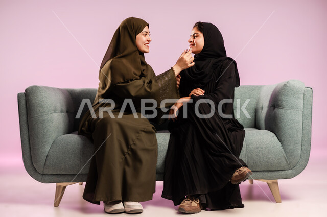 التجهز والاستعداد للذهاب مع الاصدقاء، إستخدام ادوات الميكاب تتوريال، خبيرة تجميل محترفة، بورتريه لشابة عربية خليجية سعودية ترتدي العباءة والحجاب تقوم بوضع المكياج وتزين صديقتها بتعابير وايماءات السرور، الاهتمام بالأناقة والمظهر الخارجي، خلفية ملونة