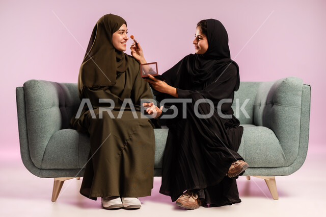Using makeup tools tutorial, getting ready to go out with friends, professional makeup artist, portrait of a young Saudi Arabian Gulf woman wearing an abaya and hijab applying makeup and adorning her friend with expressions and gestures of pleasure, interest in elegance and outward appearance, colorful background