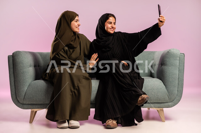 توثيق اللحظات السعيدة باستخدام الهاتف المحمول، بورتريه لإمرأة عربية خليجية سعودية ترتدي العباءة والحجاب تقوم بالتقاط صورة سيلفي تذكارية مع صديقتها بتعابير وايماءات السرور والسعادة، قضاء اوقات ممتعة برفقة الاصدقاء، جلسات وناسة نسائية، خلفية ملونة
