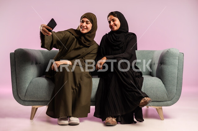 توثيق اللحظات السعيدة باستخدام الهاتف المحمول، بورتريه لإمرأة عربية خليجية سعودية ترتدي العباءة والحجاب تقوم بالتقاط صورة سيلفي تذكارية مع صديقتها بتعابير وايماءات السرور والسعادة، قضاء اوقات ممتعة برفقة الاصدقاء، جلسات وناسة نسائية، خلفية ملونة