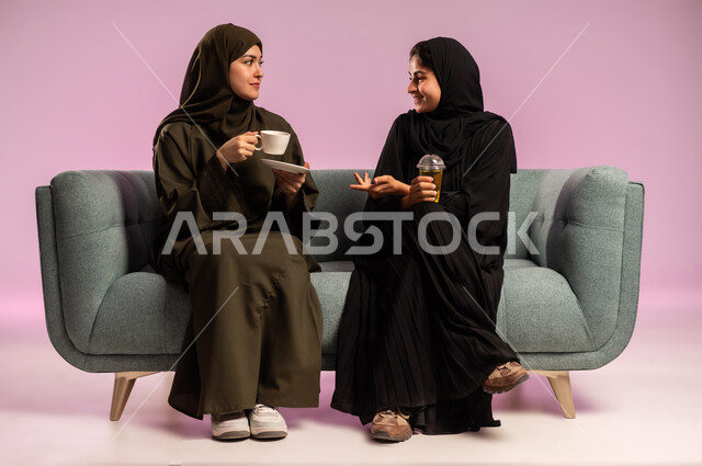 الجلوس والاستمتاع بالمشروبات الساخنة مع الاصدقاء, جلسة وناسة نسائية، قضاء الوقت باحتساء المشروب المفضل, بورتريه لشابة عربية خليجية سعودية ترتدي العباءة والحجاب تجلس على الكنبة بجانب صديقتها, تبادل الاحاديث والاخبار، خلفية ملونة