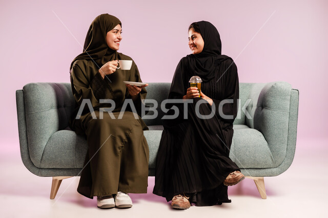 Sitting and enjoying hot drinks with friends, a women's gathering, spending time sipping their favorite drink, a portrait of a young Saudi Arabian Gulf woman wearing an abaya and hijab sitting on the couch next to her friend, exchanging conversations and news, a colorful background