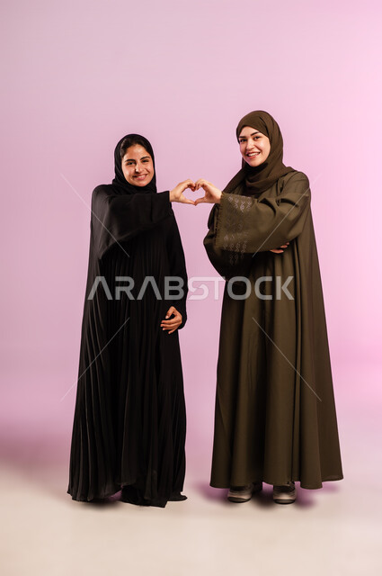 إشارة يدين على شكل قلب حب بايماءات وجه ويدين تدل على الشعور بالحب، بورتريه لإمرأتين عربيتين خليجيتين سعوديتين ترتديان العباءة والحجاب تقفان بتعابير السرور، العلاقة الايجابية الاخوية والحب والمودة، صورة الجسم بالطول الكامل، خلفية ملونة