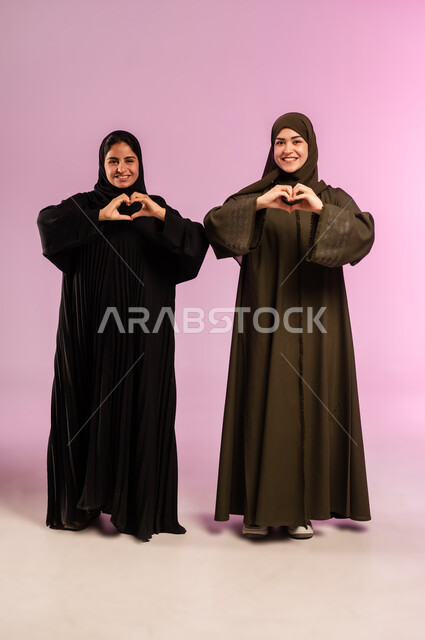إشارة يدين على شكل قلب حب بايماءات وجه ويدين تدل على الشعور بالحب، بورتريه لإمرأتين عربيتين خليجيتين سعوديتين ترتديان العباءة والحجاب تقفان بتعابير السرور، العلاقة الايجابية الاخوية والحب والمودة، صورة الجسم بالطول الكامل، خلفية ملونة