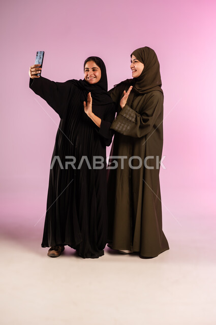 توثيق اللحظات السعيدة، استخدام الأجهزة التقنية المتطورة، بورتريه لشابة عربية خليجية سعودية محجبة ترتدي العباءة والحجاب تقوم بالتقاط صور السيلفي التذكارية مع صديقتها بإيماءات السعادة والسرور، صورة الجسم بالكامل، خلفية ملونة